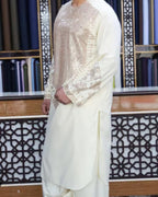 Person wearing a white outfit with intricate gold embroidery in front of a decorative wall.
