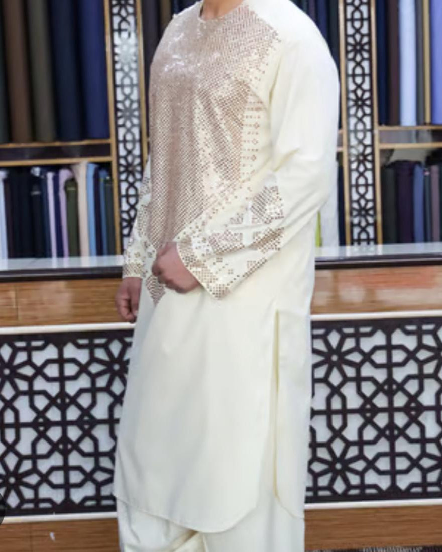 Person wearing a white outfit with intricate gold embroidery in front of a decorative wall.